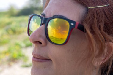 Güneş gözlüğü takan, rüzgar tarlasını yansıtan bir kadının yakın çekimi. Sürdürülebilir enerjinin ve kadınların yenilenebilir enerji sektörlerindeki rolünün, doğal bir zemine karşı belirlenmesi