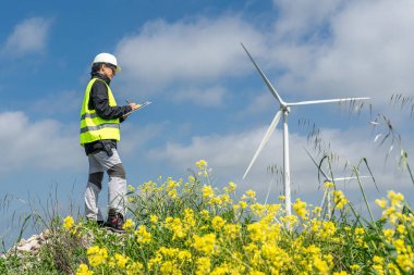 Güvenlik yeleği ve kask takan kadın mühendis, sarı çiçeklerin arasındaki rüzgar türbinlerini incelerken bir panoya notlar yazıyor ve sürdürülebilir enerjiyi destekliyor.