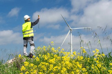 Sarı çiçekli bir alanda rüzgar hızını ölçen güvenlik yeleği ve kask takan mühendis yenilenebilir enerji için rüzgar türbini performansını değerlendiriyor.