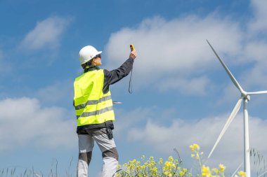 Güvenlik yeleği ve kask giyen kadın mühendis rüzgar türbinine yakın bir anemometreyle rüzgar hızını ölçüyor. Sürdürülebilir enerji üretimine ve mavi gökyüzü altında daha temiz bir çevreye katkıda bulunmak