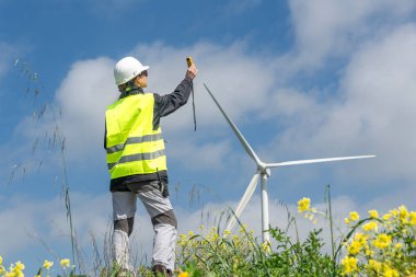 Güvenlik yeleği ve kask takan kadın mühendis anemometreyle rüzgar hızını ölçüyor, sarı çiçek tarlasında rüzgar türbininin verimliliğini hesaplıyor.