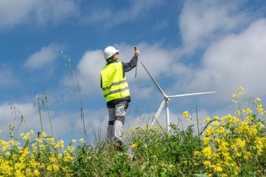 Güvenlik yeleği ve miğfer giyen kadın mühendis anemometre kullanarak rüzgar türbininin yanında rüzgar hızını ölçüyor, yenilenebilir enerji ve sürdürülebilir gelişimi teşvik ediyor.