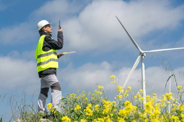 Güvenlik yeleği ve miğfer giyen kadın mühendis, belgeleri tutarken telsiz kullanıyor, güneşli bir günde sarı çiçeklerle rüzgar türbinlerinin yanında duruyor.