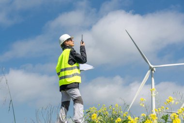 Güvenlik yeleği ve miğfer giyen kadın mühendis, mavi bulutlu bir gökyüzünün altında rüzgar türbininin yanında dokümanları tutarken telsiz kullanıyor, sürdürülebilir enerjiyi vurguluyor