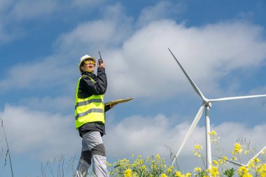 Korumalı yelek ve kask giyen kadın mühendis telsiz kullanarak ve pano tutarak sarı çiçek tarlasında rüzgar türbinlerini denetleyerek sürdürülebilir enerji çözümlerini destekliyor.