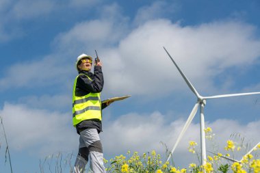 Güvenlik yeleği ve miğfer takan kadın mühendis, telsiz kullanıyor ve elinde bir pano var. Rüzgâr türbinlerinin yanında duruyor. Önplanda sarı çiçekler olan bulutlu mavi bir gökyüzünün altında.