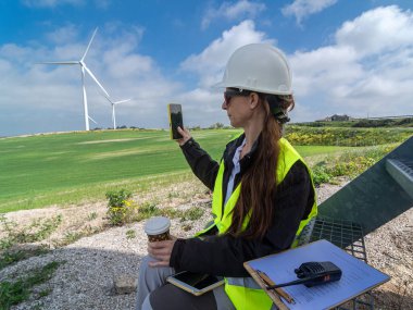 Sert şapka ve güvenlik yeleği giyen kadın mühendis, geniş yeşil alandaki rüzgar türbinlerini izlemek için akıllı telefon kullanıyor ve yenilenebilir enerji endüstrisindeki kadınların rolünü vurguluyor.
