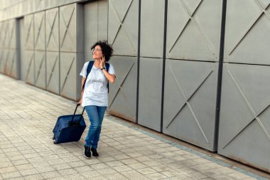 Dişi dijital göçebe, akıllı telefonuyla sohbet ederken kaldırımda yuvarlanan bavullarıyla geziyor. Şehir ortamında şık, modern bir binanın önünden geçiyor.