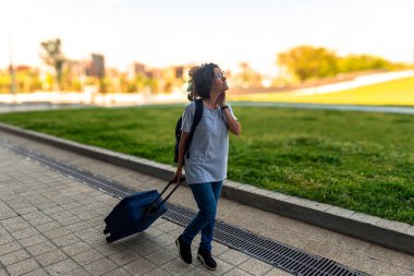 Kadın dijital göçebe, valizi ve sırt çantasıyla şehir parkında gezerken, uzaktan kumandalı çalışmanın özgürlüğünün ve esnekliğinin tadını çıkarıyor.