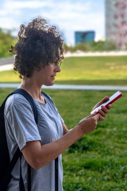 Kıvırcık saçlı ve gözlüklü dijital göçebe, sırt çantası taşıyor, akıllı telefonunu modern bir şehir parkında kullanıyor, uzaktan çalışma, seyahat ve kadınların güçlendirme kavramını somutlaştırıyor.