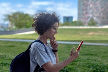 Kıvırcık saçlı ve gözlüklü düşünceli bir kadın dijital göçebe. Sırt çantası taşıyor. Modern bir şehir parkında akıllı telefonunu kullanıyor. Seyahat ruhunu somutlaştırıyor. Uzaktan kumanda. Ve kadınların güçlenmesi.