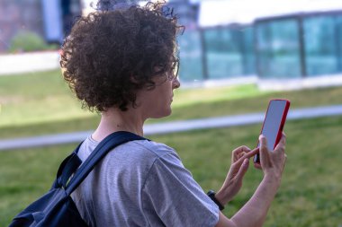 Kıvırcık saçlı, gözlük takan ve sırt çantası taşıyan dijital göçebe kadın açık havada akıllı telefon kullanıyor, muhtemelen uzaktan çalışıyor ya da seyahatleri sırasında yol gösteriyor.