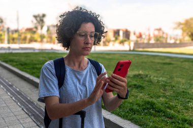 Navigasyon için akıllı telefon kullanan kadın dijital göçebe, bir şehir parkında bavulla seyahat ederken konaklama veya ulaşım rezervasyonu