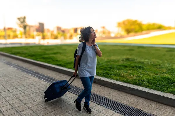 Kadın dijital göçebe, valizi ve sırt çantasıyla şehir parkında gezerken, uzaktan kumandalı çalışmanın özgürlüğünün ve esnekliğinin tadını çıkarıyor.