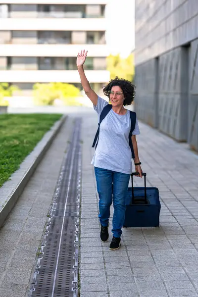 Mutlu kadın dijital göçebe, şehir ortamında çanta ve sırt çantasıyla dolaşırken, özgürlüğünün ve uzak çalışma hayatının tadını çıkarırken el sallıyor.