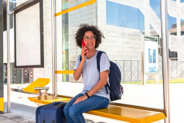 Sırt çantası ve tramvay valizli neşeli kadın dijital göçebe modern bir otobüs durağında oturmuş, ulaşımını beklerken bir telefon görüşmesi yapıyor.