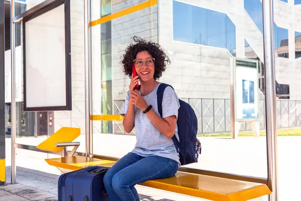 Sırt çantası ve bavuluyla mutlu bir kadın dijital göçebe modern bir otobüs durağında telefon ederken gülümsüyor, seyahatleri sırasında iş hayatının dengesinin keyfini çıkarıyor.
