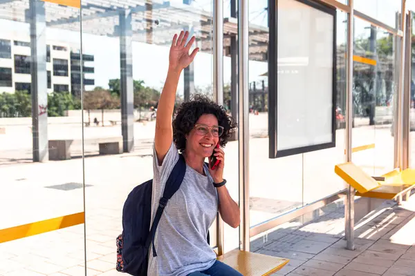 Sırt çantalı gülümseyen kadın turist, güneşli bir şehir merkezinde modern bir otobüs durağında akıllı telefonuyla sohbet ederken gelen bir otobüse coşkuyla el sallıyor.