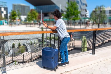 Bavuluyla seyahat ederken akıllı telefon kullanan genç bir kadın, modern bir şehir ortamında uzaktan çalışmanın özgürlüğünün ve esnekliğinin tadını çıkarıyor.