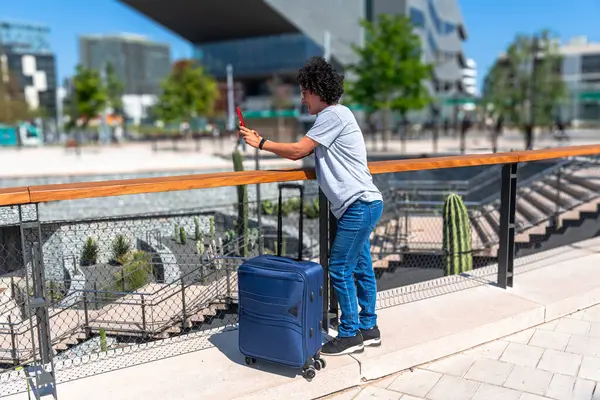 Genç bir kadın akıllı telefonuyla fotoğraf çekiyor, modern bir şehirde valiziyle seyahat ederken, dijital göçebe yaşam tarzının tadını çıkarıyor ve yeni kentsel alanlar keşfediyor.