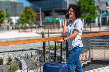 Dişi dijital göçebe canlı, modern bir şehirde yürüyor, akıllı telefonuyla sohbet ediyor, uzaktan çalışma özgürlüğünü kucaklıyor ve güneşli bir yaz gününün tadını çıkarıyor.