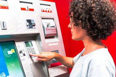 Genç kıvırcık saçlı kadın yabancı bir ülkede ATM kullanıyor, seyahat masrafları için para çekiyor, kadınların yetkilerini ve finansal bağımsızlığını sergiliyor.