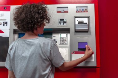 Genç kıvırcık saçlı bir kadın, para çekmek için tren istasyonundaki bir bankamatiğe mor bir kredi kartı sokuyor. Ekonomik bağımsızlık ve seyahat fırsatlarına odaklanıyor.