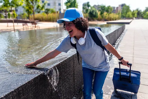 Sırt çantası ve kulaklık takan genç bir kadın, seyahat deneyiminin tadını çıkarıyor, tramvay valizini çekerken şehir çeşmesinde suya dokunuyor.