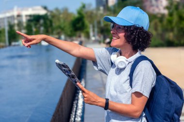 Sırt çantası ve kulaklıklı neşeli turist elinde bir harita tutarken gülümsüyor ve bir şehir parkının simgesini işaret ediyor, kadınların gücünü kucaklayan dijital bir göçebe olarak seyahat deneyiminin tadını çıkarıyor.