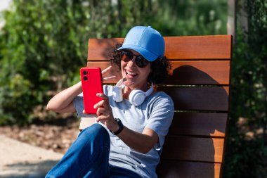Güneş gözlüğü takan ve mavi şapkalı mutlu bir kadın güneşli bir parkta bankta oturup dijital göçebe hayatı dengesi kurarken kırmızı bir akıllı telefonla video görüşmesi yapıyor.