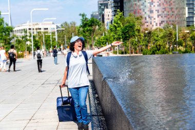 Sırt çantası ve kulaklıklı neşeli bayan turist güneşli bir yaz gününde modern bir şehirde çantalarını fıskiyenin yanına çekiyor ve İspanya 'nın Barselona kentinde seyahat deneyiminin tadını çıkarıyor.