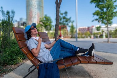 Genç serbest çalışan gülüyor, uzaktan çalışmaya ara veriyor, Barselona parkında modern bir bankta dinleniyor, dijital göçebe yaşam tarzını benimsiyor ve sağlıklı bir iş hayatı dengesi kuruyor.