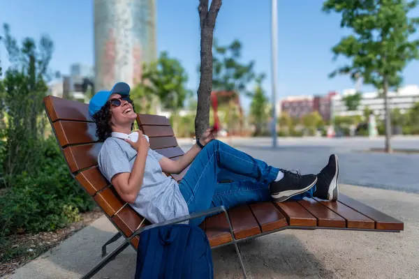 Genç serbest çalışan gülüyor, uzaktan çalışmaya ara veriyor, Barselona parkında modern bir bankta dinleniyor, dijital göçebe yaşam tarzını benimsiyor ve sağlıklı bir iş hayatı dengesi kuruyor.