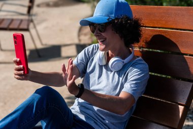 Mavi şapkalı ve kulaklıklı mutlu bir kadın güneşli bir parkta, ahşap bir bankta oturup uzaktan çalışmanın keyfini çıkarırken akıllı telefonuyla sesleniyor.