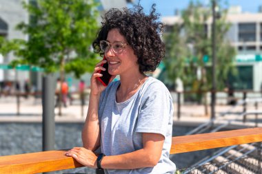 Kıvırcık saçlı ve gözlüklü neşeli serbest yazar, modern şehir ortamında ahşap korkuluklara yaslanıp dijital göçebe yaşam tarzını benimserken bir telefon görüşmesi yapıyor.