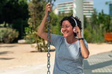 Güler yüzlü dijital göçebe kablosuz kulaklık takıyor güneşli bir şehir parkında dizüstü bilgisayarıyla çalışıyor, bir salıncak zinciri tutuyor ve esnek iş yaşam tarzının keyfini çıkarıyor.