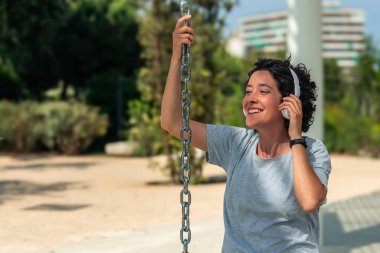 Gülümseyen dijital göçebe, bir şehir parkında durup salıncak zinciri tutarken kulaklıkla müzik dinliyor ve uzaktan çalışma özgürlüğünün tadını çıkarıyor.