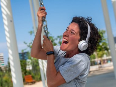 Kıvırcık saçlı, kulaklık takan ve akıllı bakışlı neşeli kadın, modern bir şehir parkında zincir sallayarak boş zamanının tadını çıkarıyor, neşe ve özgürlüğü ifade ediyor.
