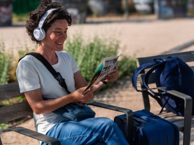 Mutlu kadın dijital göçebe kulaklık takıyor ve broşür okuyor. Sırt çantası ve valiziyle parkta bankta oturuyor, dışarıda iş hayatının tadını çıkarıyor.