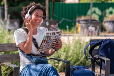 Mutlu kadın dijital göçebe kulaklık takıyor, müzik dinliyor, parktaki bankta fotoğraf albümüne göz atıyor, bavulları yanında, seyahat ve uzaktan çalışmayı kucaklıyor.