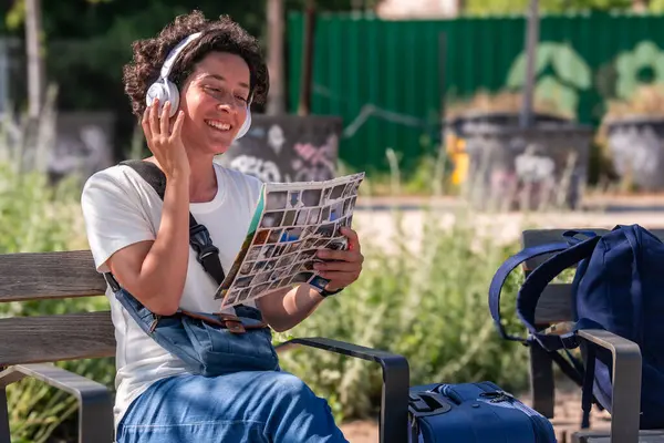 Mutlu kadın dijital göçebe kulaklık takıyor, müzik dinliyor, parktaki bankta fotoğraf albümüne göz atıyor, bavulları yanında, seyahat ve uzaktan çalışmayı kucaklıyor.