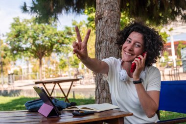 Mutlu kadın serbest yazar, cep telefonuyla konuşurken, tablet ve hesap makinesiyle parkta çalışırken, esnek iş ve özgürlüğün tadını çıkarırken zafer işareti yapıyor.