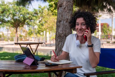 Kadın serbest çalışan gülümseyen ve cep telefonuyla konuşan, not alan ve şehir parkında tablet kullanan, iş hayatı dengesinin keyfini çıkaran ve dijital göçebe yaşam tarzını benimseyen biri.