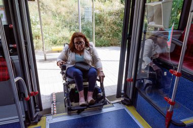 Motorlu tekerlekli sandalye kullanan bir kadın toplu taşıma otobüsüne giriş rampasından giriyor ve engelli bireyler için erişilebilirlik gösteriyor. Katılım kavramı, uyarlanmış toplu taşıma