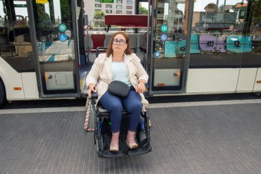 Tekerlekli sandalye kullanan bir kadın modern bir toplu taşıma otobüsünden iniyor, yerleşik bir giriş rampası kullanıyor, şehir ulaşımında erişilebilirliği gösteriyor. Katılım kavramı, uyarlanmış toplu taşıma