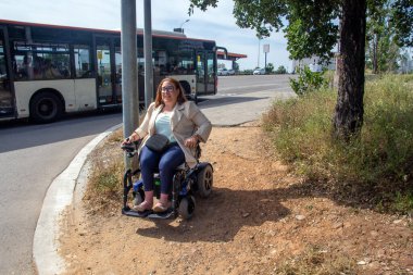 Smiling woman sitting in an electric wheelchair on a dirt patch by a bus stop, waiting for public transport on a sunny day. Concept of inclusion, adapted public transport