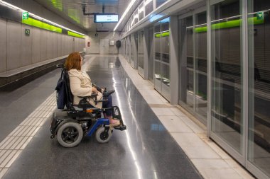 Motorlu tekerlekli sandalye kullanan bir kadın modern bir metro istasyonunda bekliyor, toplu taşımacılıkta ulaşılabilirliğini gösteriyor. Katılım kavramı, uyarlanmış toplu taşıma