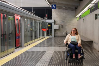 40 'lı yaşlarda beyaz bir iş kadını, metro trenini beklerken tekerlekli sandalye kullanıyor, ulaşılabilir toplu taşıma araçlarının keyfini çıkarıyor. Katılım kavramı, uyarlanmış toplu taşıma