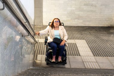Tekerlekli sandalyesiyle metro istasyonunda gezinen iş kadını, destek için ulaşılabilir rampa ve tırabzanları kullanıyor, hareketlilik ve kapsamı ön plana çıkarıyor. Katılım kavramı, uyarlanmış toplu taşıma