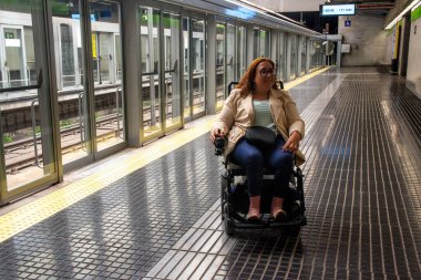 Tekerlekli sandalye kullanan genç bir kadın, ulaşılabilir bir metro platformunda, dokunsal kaldırımları ve peron kapıları olan trenin gelişi için bekliyor. Katılım kavramı, uyarlanmış toplu taşıma